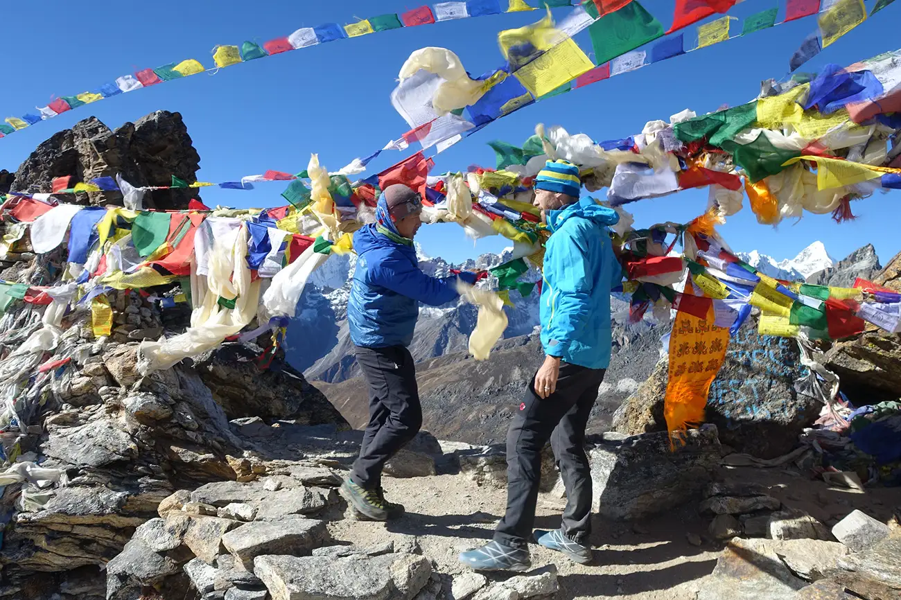 Two hikers exchanging a playful high-five under colorful prayer flags at the summit of Renjo La Pass with snow-capped mountains in the background.