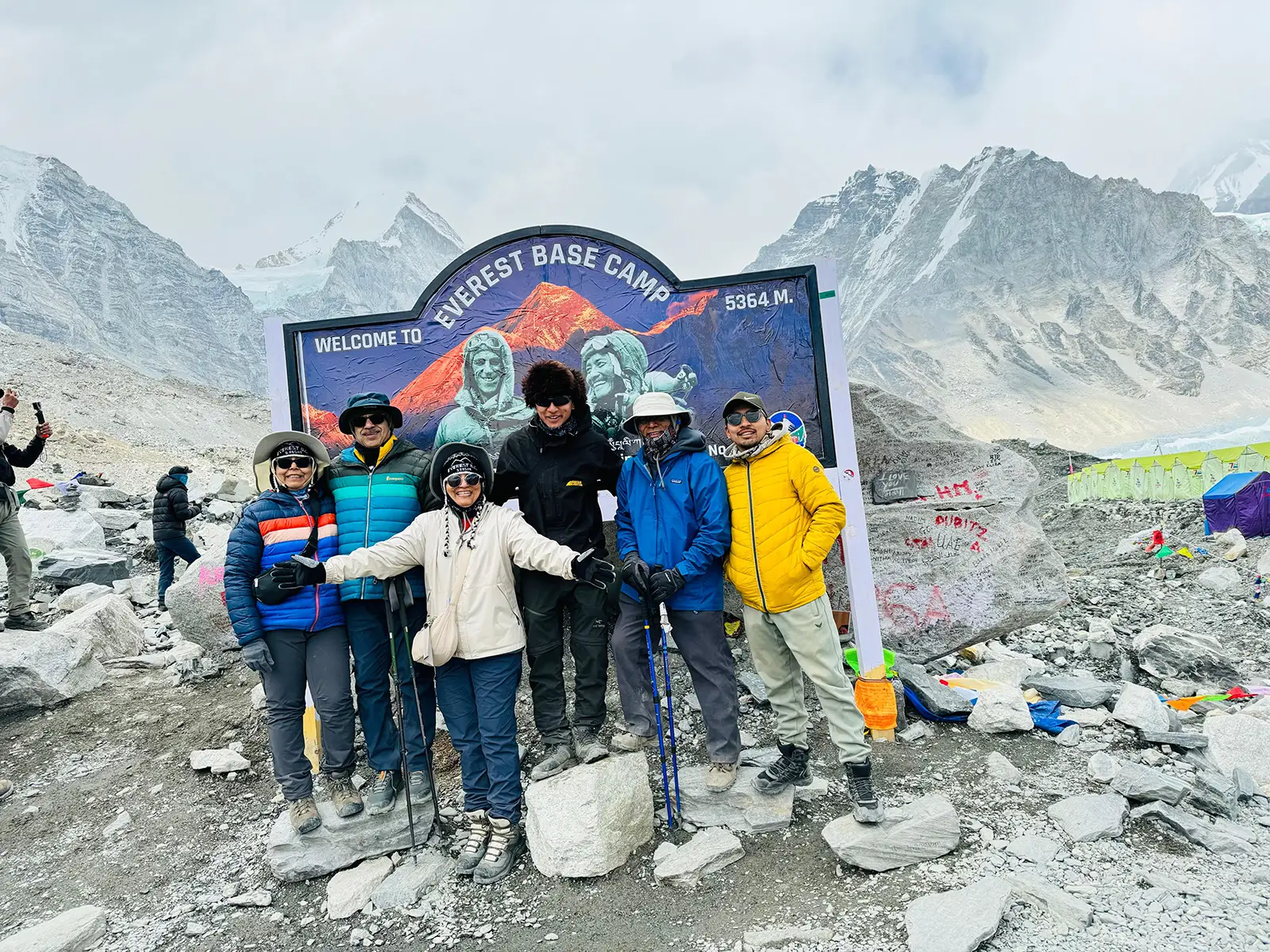 A group of trekkers celebrating at Everest Base Camp, with the iconic prayer flags fluttering in the background and the snow-capped peak of Mount Everest visible in the distance.
