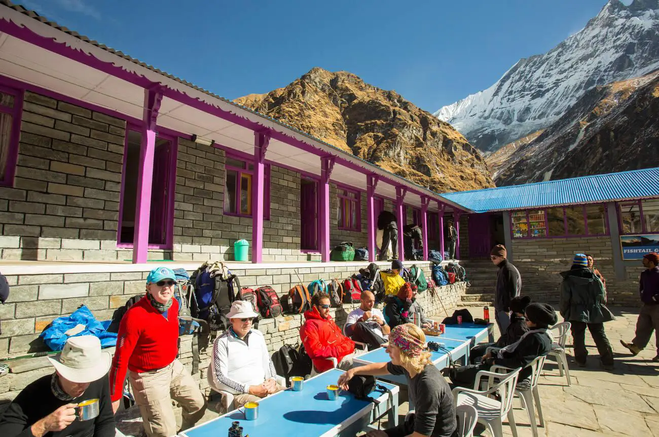 Trekkers relaxing at a tea house lodge at Machapuchare Base Camp, Nepal, surrounded by Himalayan mountains.