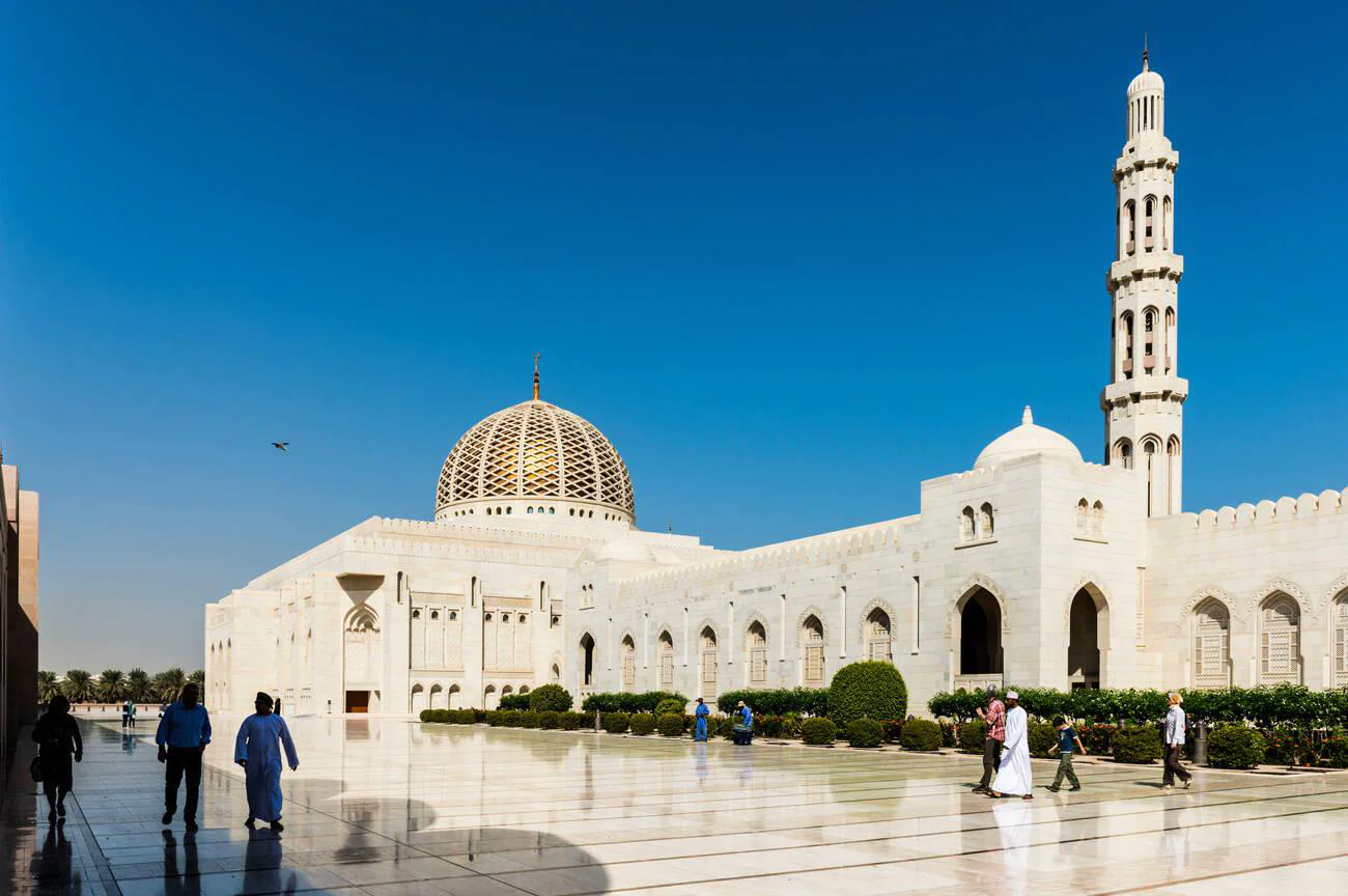 Visitors walking in the vast courtyard of Sultan Qaboos Grand Mosque under the clear blue sky in Muscat, Oman.