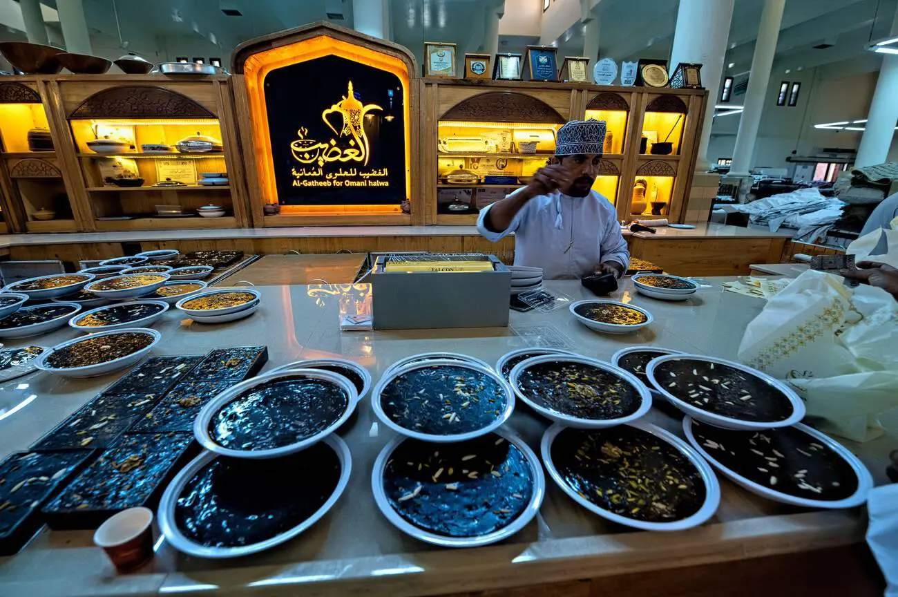 Traditional Omani halwa displayed for sale in a shop in Nizwa, Oman, with a shopkeeper interacting with customers.