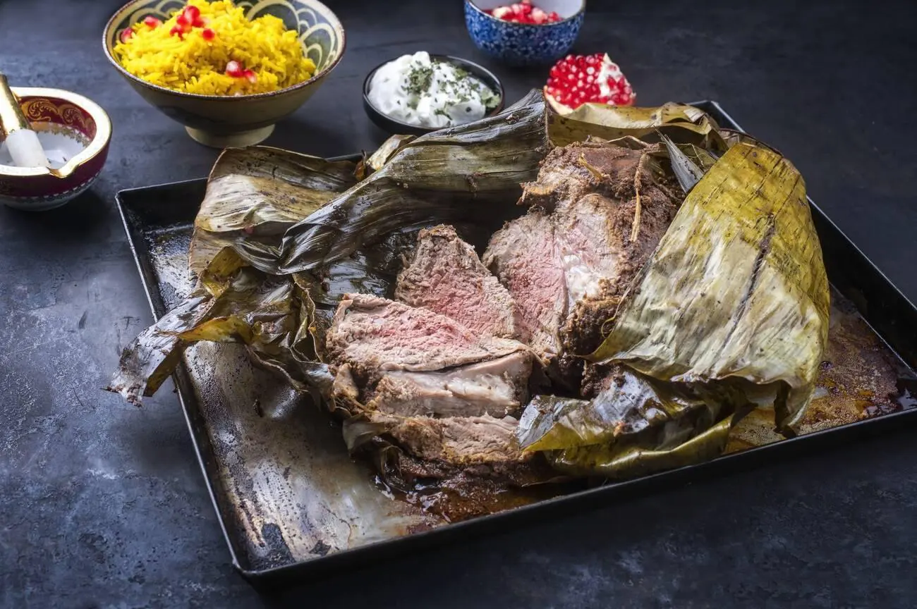 A traditional Omani lamb shuwa, slow-cooked and wrapped in banana leaves, served with saffron rice and yogurt on a dark surface.