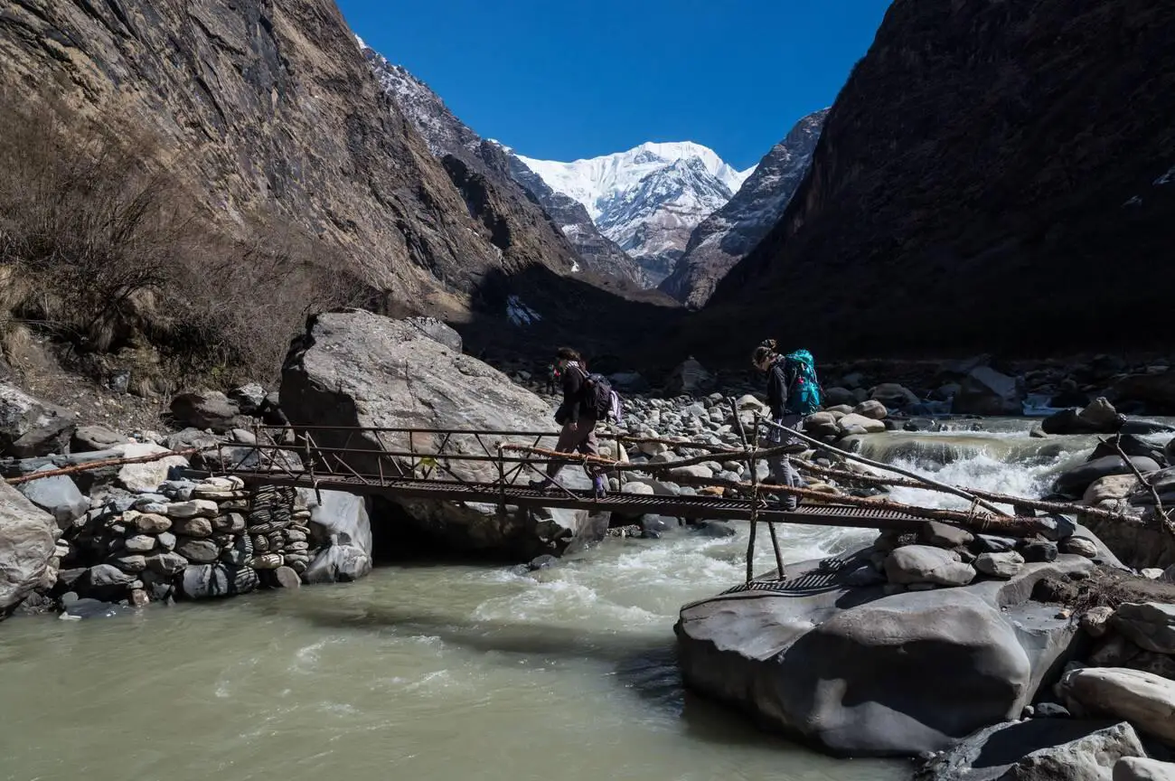 Trekkers crossing a bridge on the way to Machapuchare Base Camp in Nepal with snow-capped mountains in the background.