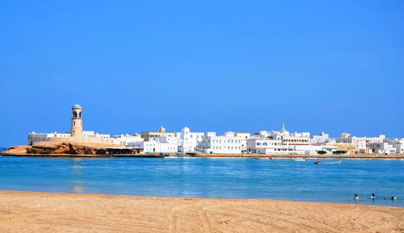 Coastal view of Sur, a historic city in Oman, with white-washed buildings and a lighthouse overlooking the calm blue sea.