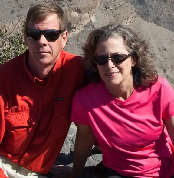 A middle-aged couple posing on the rocky cliffs of Jebel Shams, Oman, with a steep canyon in the background.