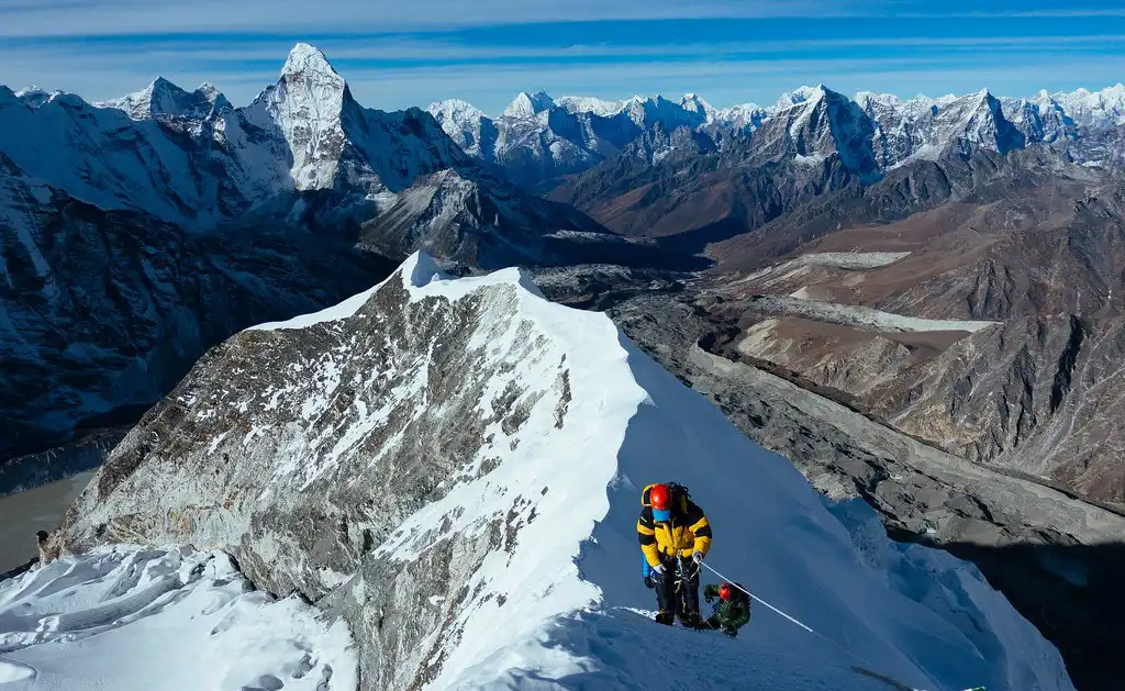 Climber nearing the summit of Island Peak in Nepal, ascending a narrow, snow-covered ridge with breathtaking views of the surrounding Himalayan mountains.