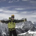 Climber standing triumphantly at the summit of Island Peak with ice axe raised, surrounded by snow-capped Himalayan mountains.
