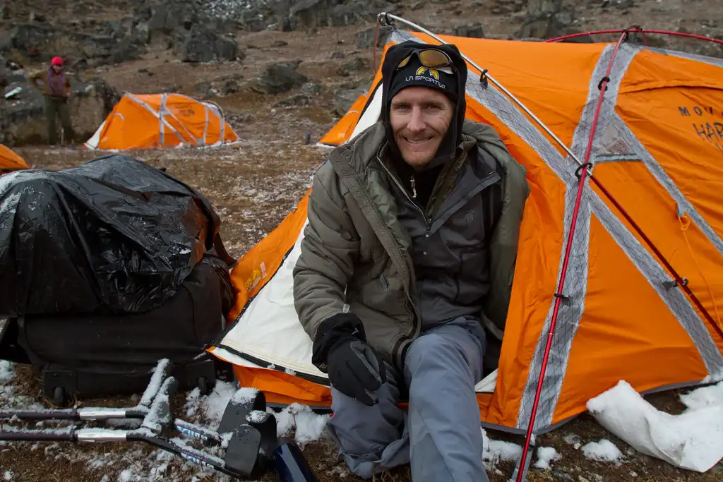 A climber sits outside an orange tent at Lobuche Base Camp, wearing a beanie and jacket, smiling as they prepare for the ascent in the cold, rugged environment.