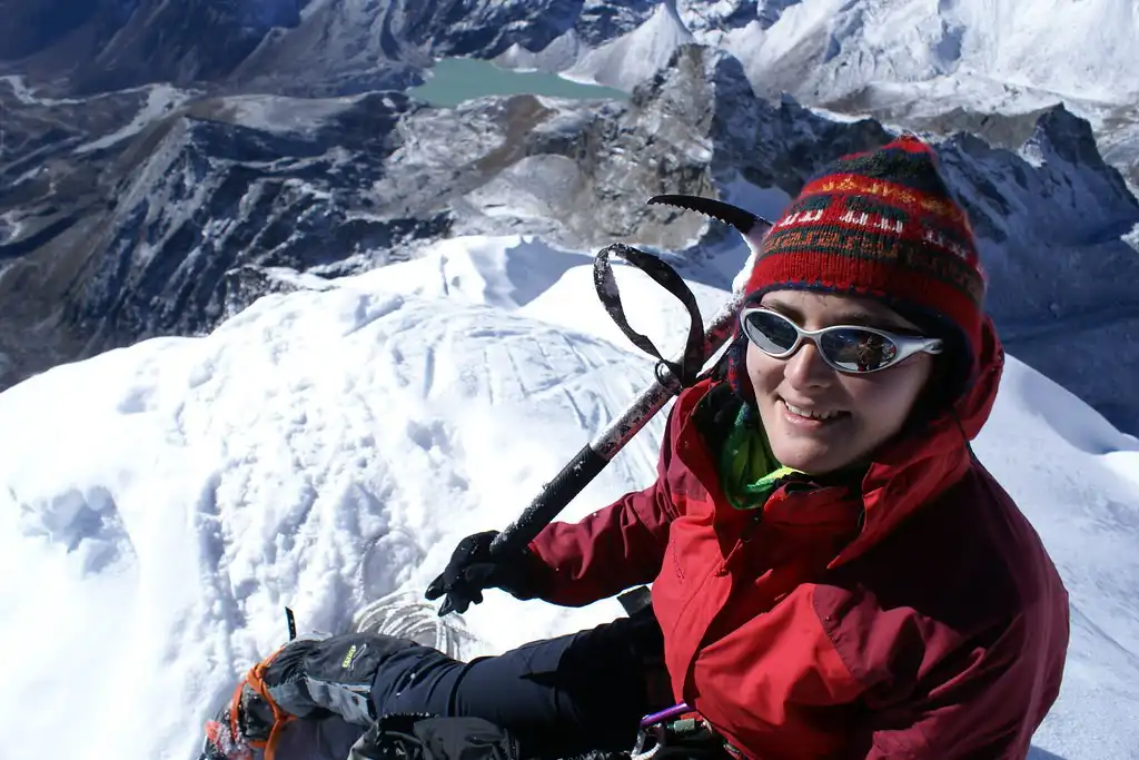 A climber wearing a red jacket and woolen hat smiles at the camera while holding an ice axe on the snowy summit of Lobuche Peak, with a mountain valley below.