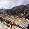 Trekkers on a rugged trail overlooking Dingboche village in the Khumbu Valley, with terraced fields and snow-capped peaks in the background.