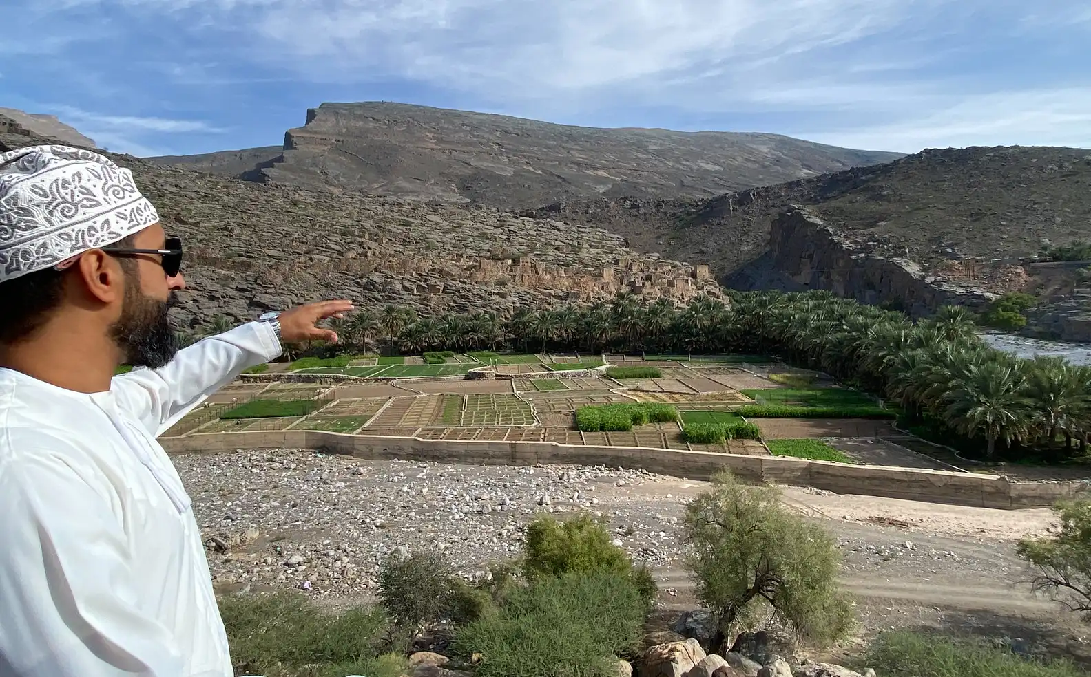 Man in traditional Omani attire pointing towards lush green terraces in Wadi Ghul, Oman, with rugged mountains and a valley in the background.