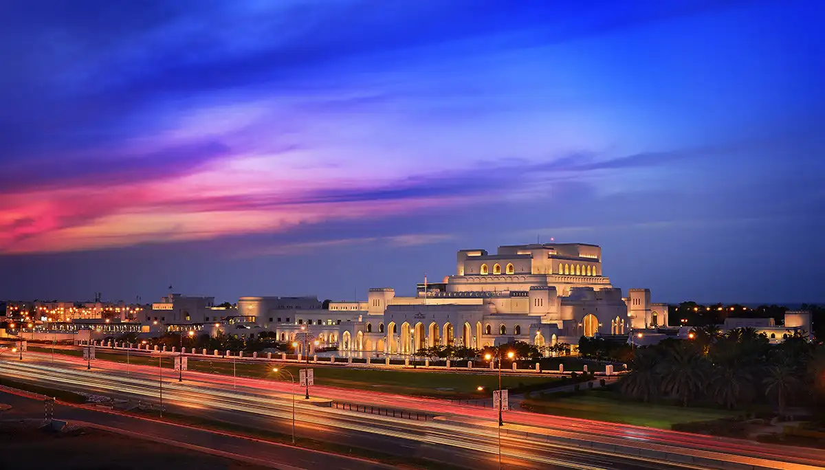 The Royal Opera House in Muscat, Oman, beautifully lit up against a colorful evening sky with streaks of vibrant blue and pink.