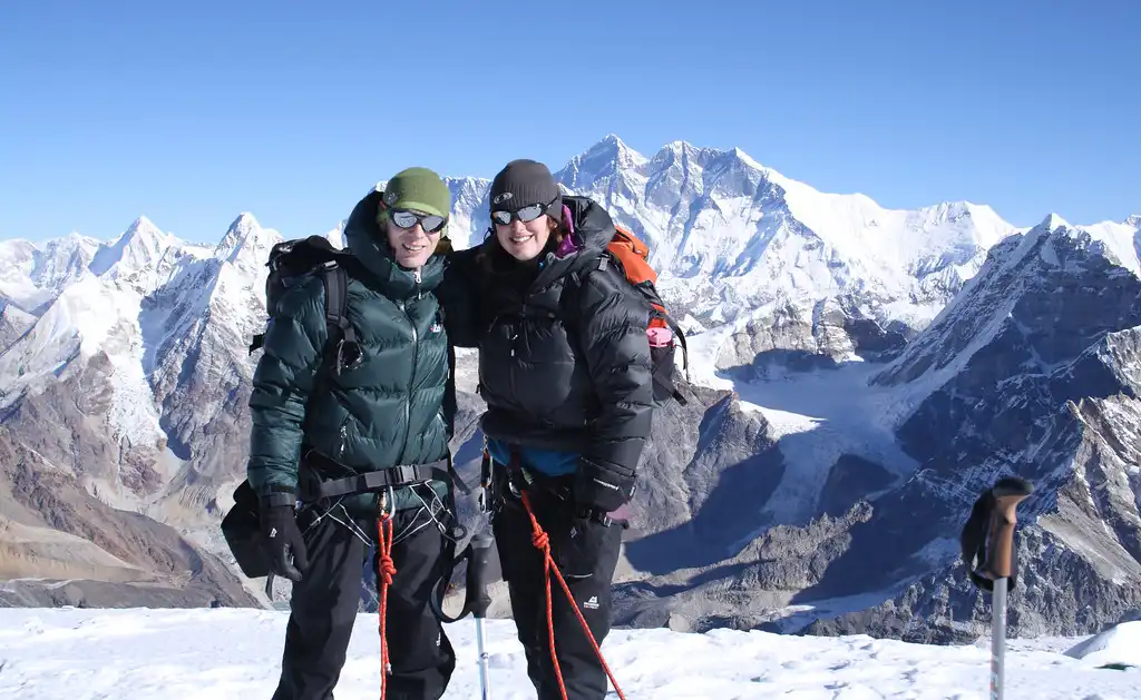 Twee klimmers in hun warme bergbeklimmerskleding staan ​​lachend op de top van Mera Peak, met de Himalaya op de achtergrond.