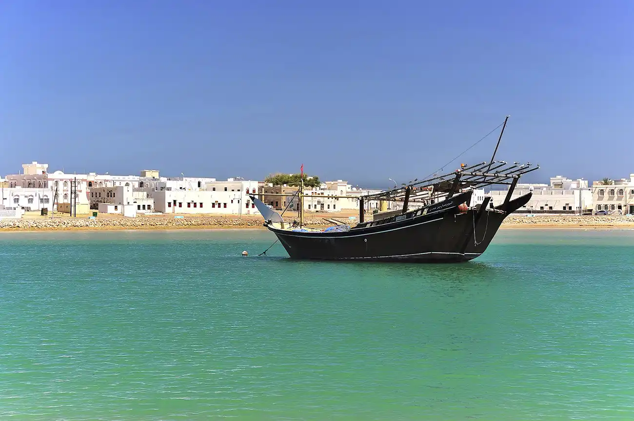 Traditional dhow boat anchored in the turquoise waters of Sur’s lagoon, with white buildings in the background in Oman.