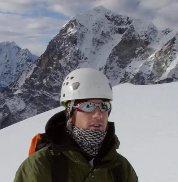A mountaineer in a green jacket and helmet stands on the snow-covered slopes of Lobuche, with a vast panorama of the Himalayan mountain range in the background.