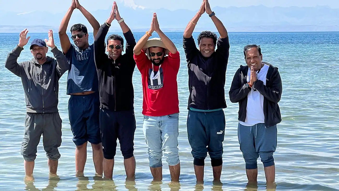 A group of devotees standing in the shallow waters of Mansarovar Lake, with hands joined in prayer or raised in a gesture of reverence, set against the backdrop of clear blue water and distant mountains.