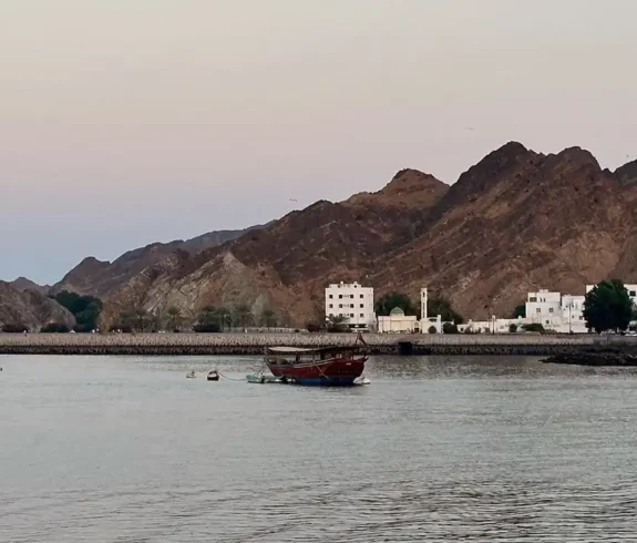 Panoramic view of Mutrah Corniche at dusk with traditional dhow boats and rugged mountains in the background.