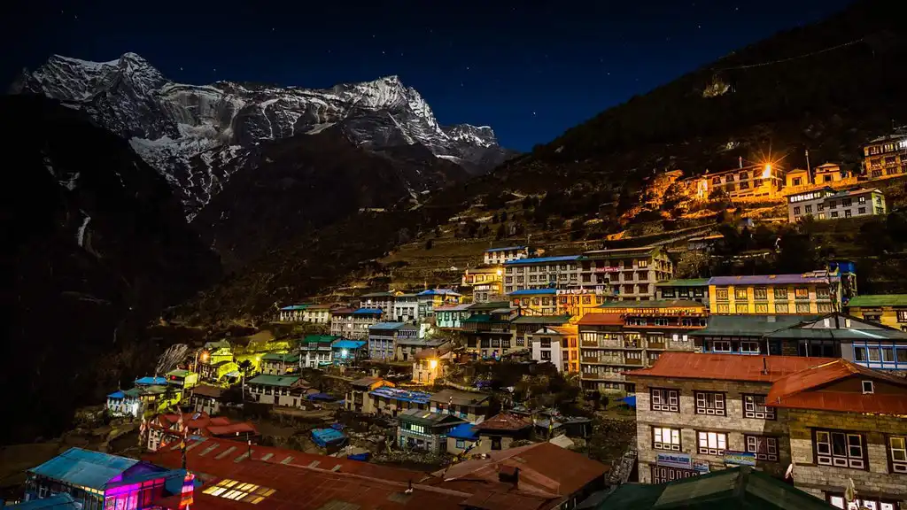 Night view of Namche Bazaar brightly lit with colorful lights under a starry sky, nestled in the mountainous landscape of Sagarmatha National Park.