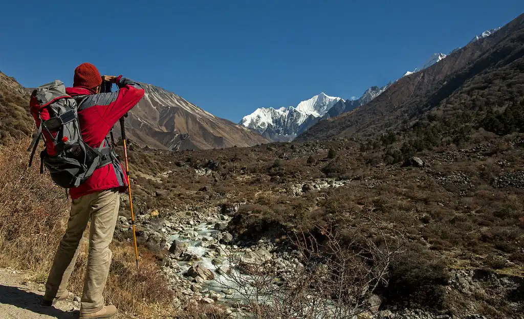 A photographer in a red jacket with a backpack stands on a trail in Langtang National Park, Nepal, capturing the snow-capped mountain peaks with his camera.