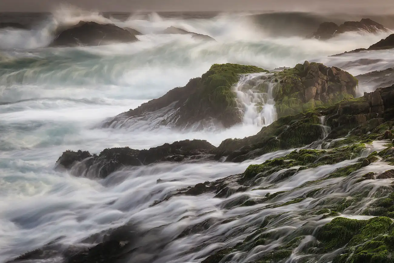 Waves crashing against moss-covered rocks near Salalah, Oman, creating a misty, dramatic scene.