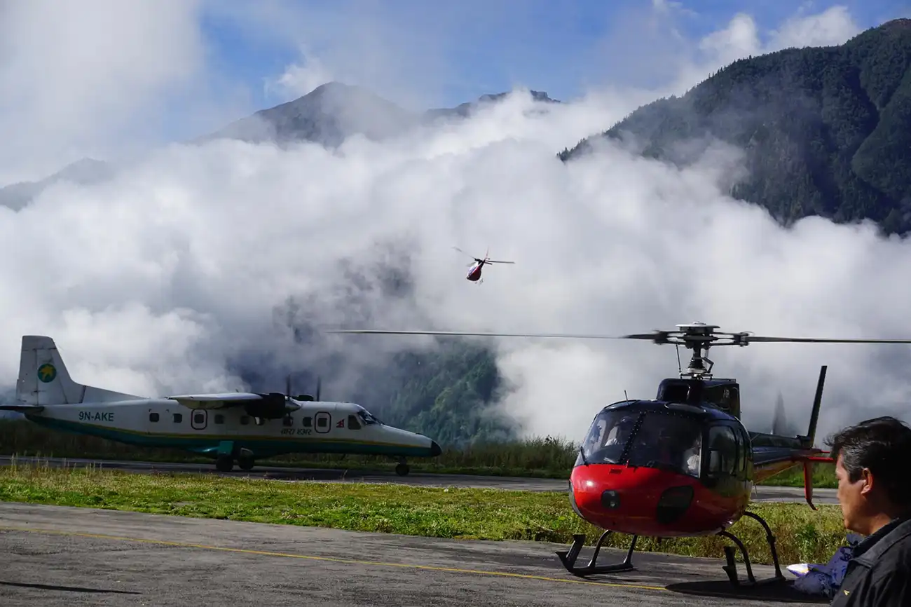Een klein vliegtuig en helikopters op een landingsbaan in de bergen. Eén helikopter stijgt op, omgeven door mistige wolken en weelderige groene heuvels.