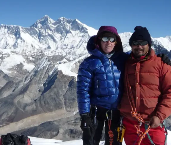 Two climbers in high-altitude climbing gear pose together at the summit of Mera Peak, with a panoramic view of the Himalayas in the background.