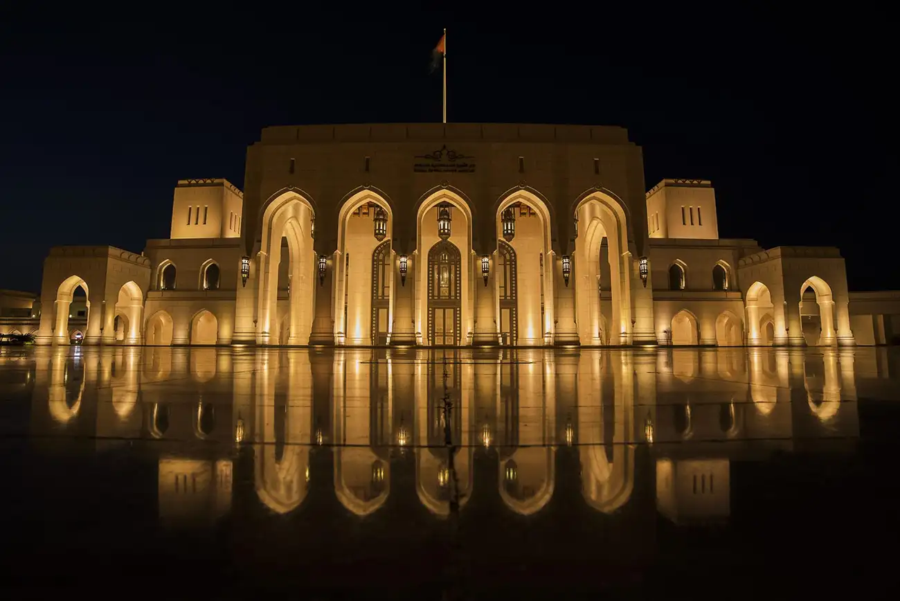 The Royal Opera House in Muscat, Oman, illuminated at night with golden lighting, reflecting beautifully on the polished courtyard.