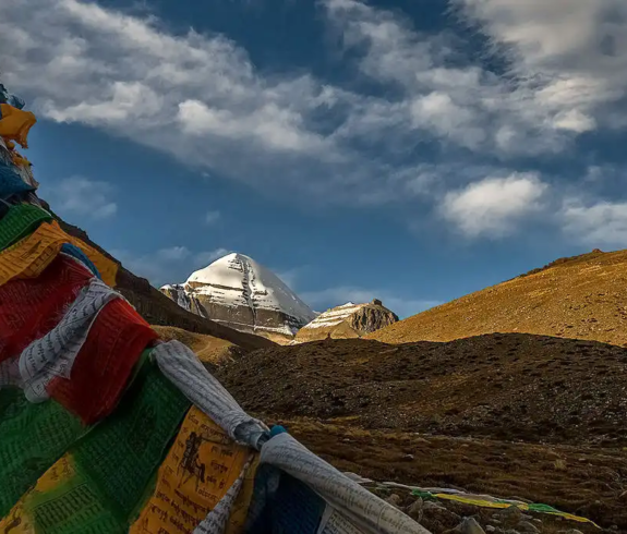 Mount Kailash rises beyond colorful prayer flags at sunrise, with golden hills, dark clouds, and sacred Tibetan plateau scenery.