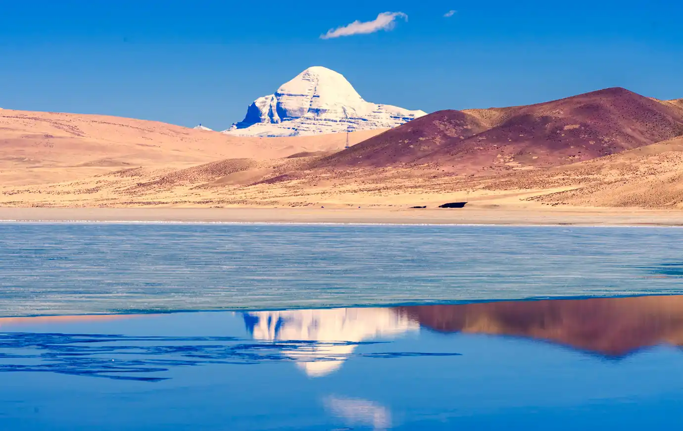 Mount Kailash reflects on the calm waters of Lake Mansarovar at sunrise, surrounded by golden Tibetan plateau hills and clear blue sky.