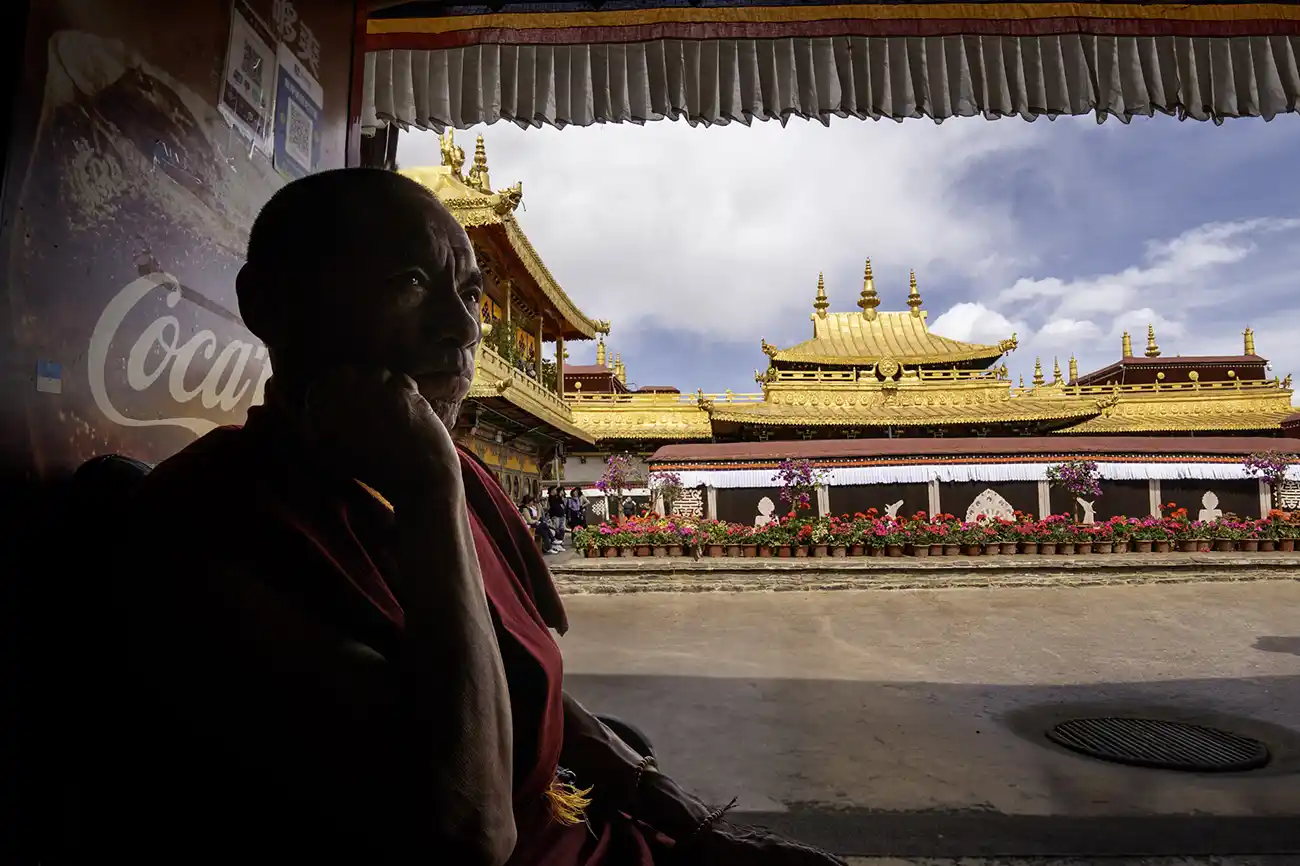 A monk in contemplation inside the Jokhang Temple, with the golden roofs of the temple visible in the background.