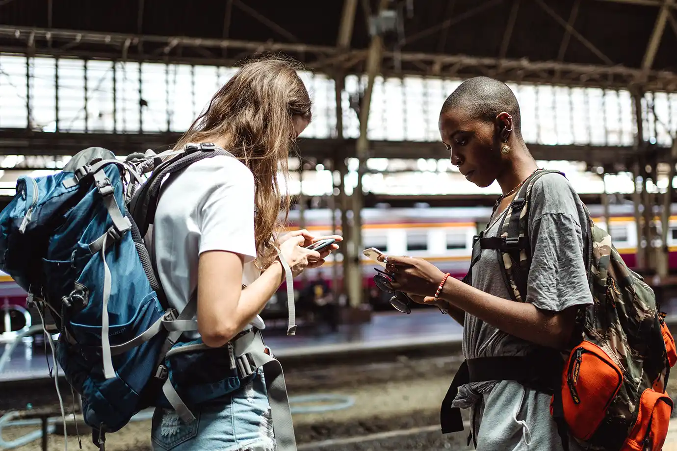 Two young women carrying large backpacks standing side by side while checking their smartphones at a covered railway station platform.