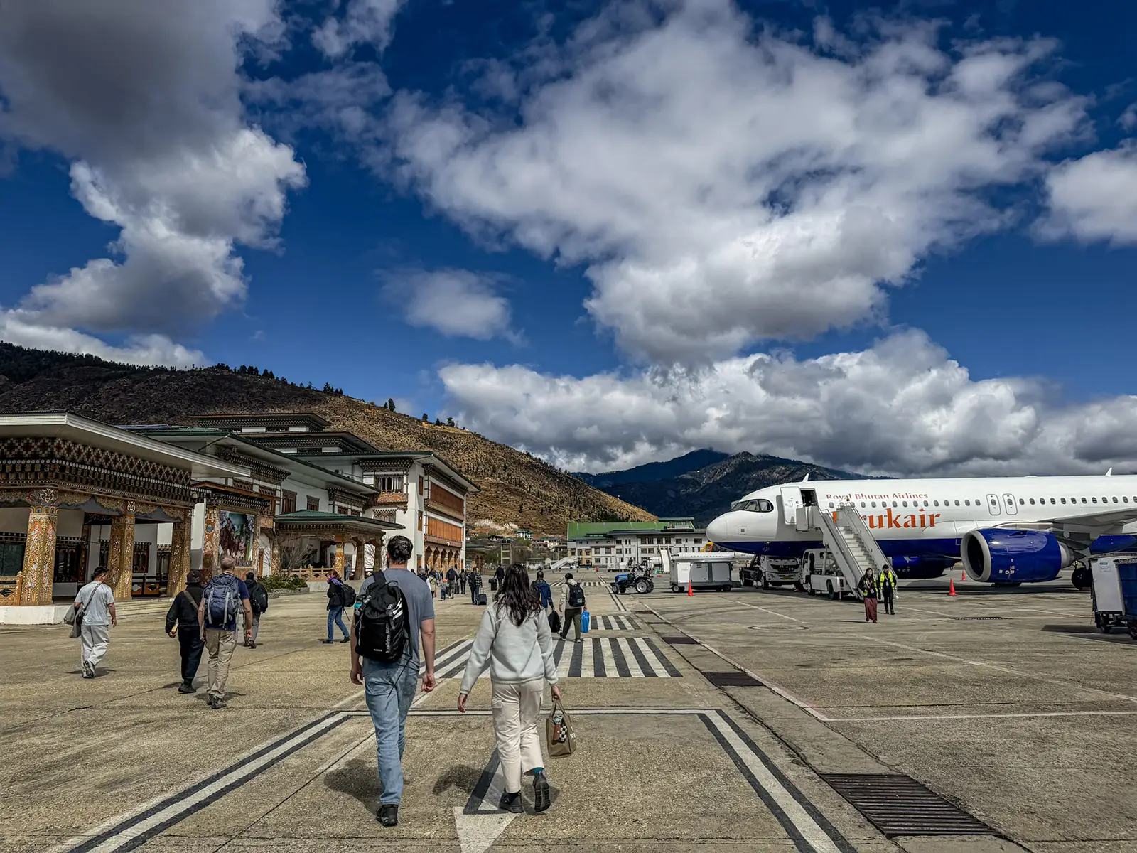Travelers walking across tarmac toward traditional Bhutanese airport terminal with Drukair aircraft