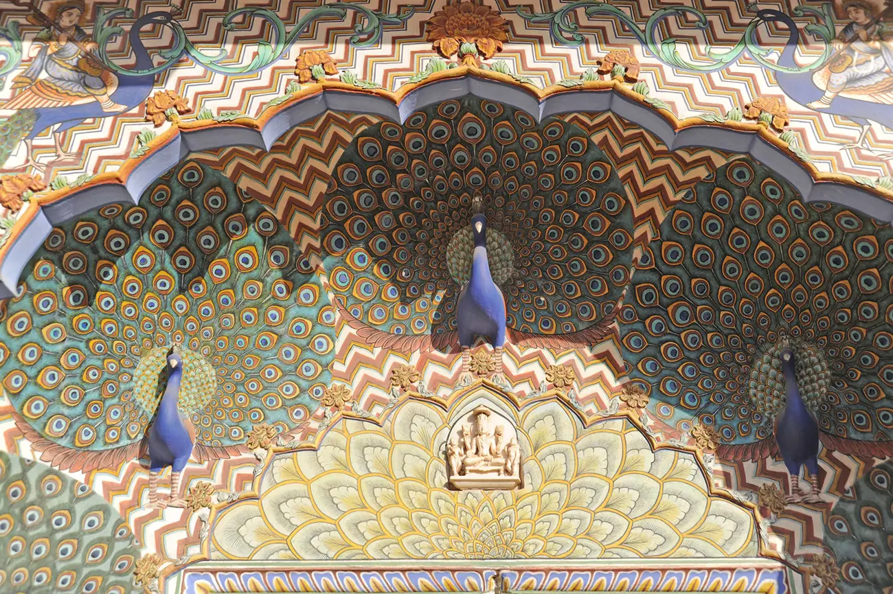 A close-up of a vibrantly painted archway in the Chandra Mahal Museum, featuring three peacocks with their feathers fanned out, forming the pattern of the arch. The colors are predominantly blues, greens, and browns, with intricate chevron and floral patterns surrounding the peacocks.