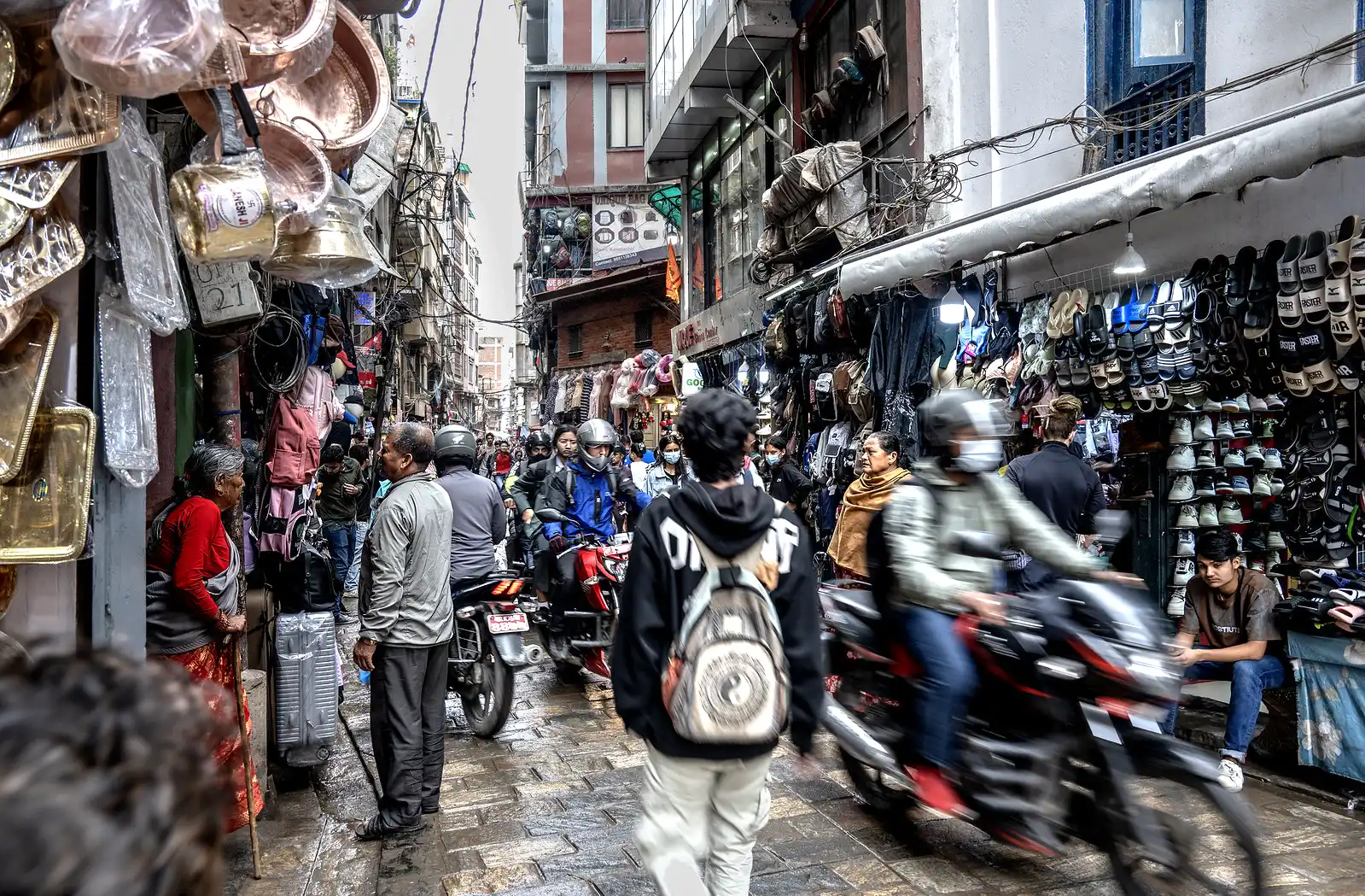 Pedestrians and motorcycles navigate a busy, narrow stone street lined with copper shops and shoe vendors in Ason Bazaar, Kathmandu.
