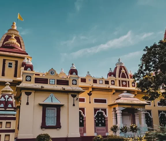 The magnificent Birla Mandir, a large cream and red Hindu temple with intricate spires, stands majestically amidst lush green gardens under a vibrant blue sky with scattered clouds.
