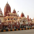 The vibrant Birla Temple in New Delhi, featuring distinct red and cream spires and domes, stands beside a busy road with a black fence, under a bright, clear sky.