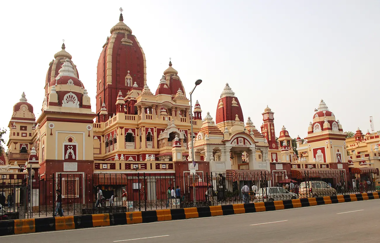 The ornate Shri Laxmi Narayan Temple (Birla Mandir) in Delhi, with its red and yellow spires, seen behind a white marble pavilion situated on a green lawn under a clear blue sky.