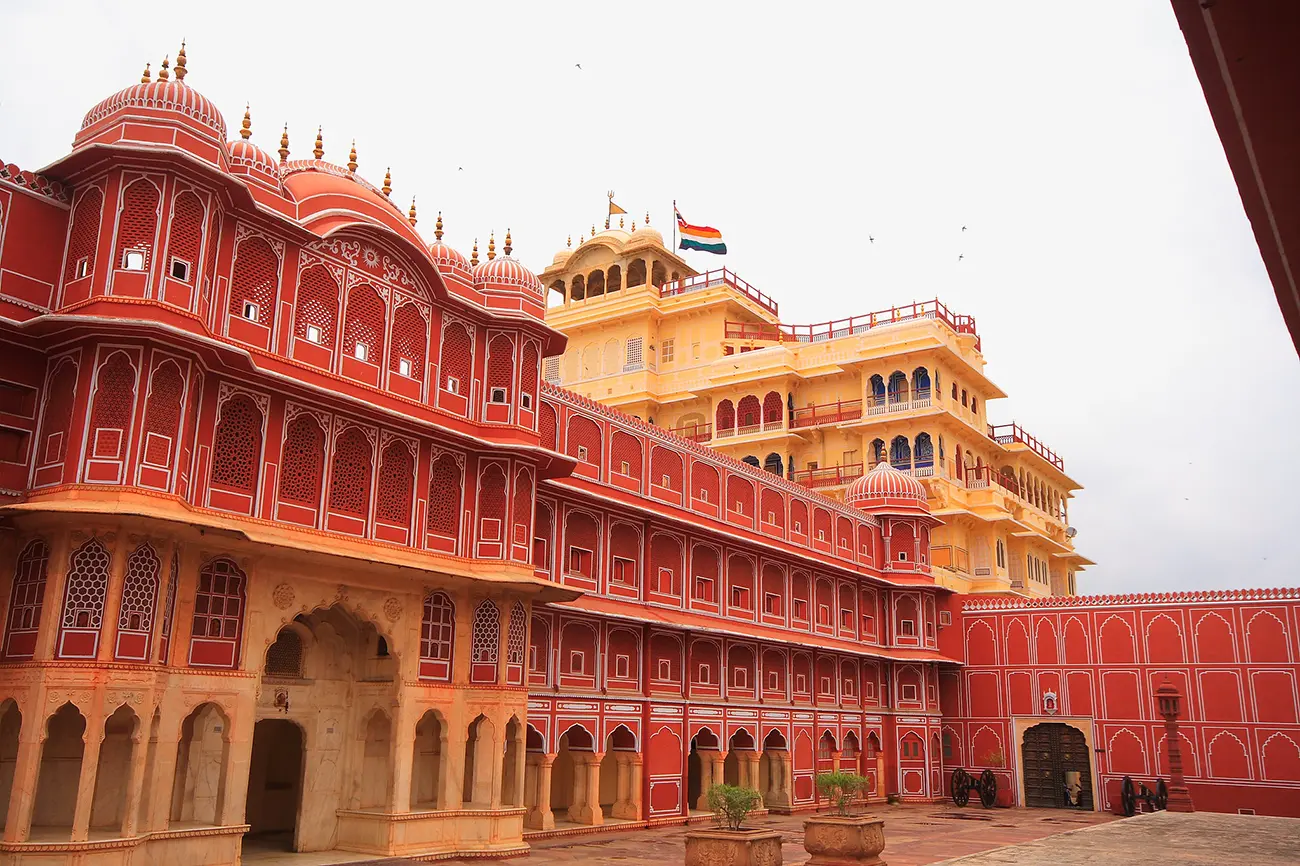 The vibrant red and cream-colored facades of Chandra Mahal in Jaipur, with multiple arched windows, balconies, and domed rooftops under a pale sky. An Indian flag flies from the highest point of the cream-colored building.