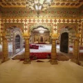A richly decorated room in the City Palace Museum, with ornate golden pillars and arches adorned with red and green gemstone-like details, leading to an inner chamber with red seating and white floor coverings.