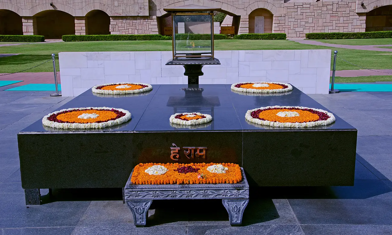 The black marble memorial stone at Raj Ghat, adorned with flower petals, with the eternal flame burning above, and the inscription "He Ram" in Hindi.