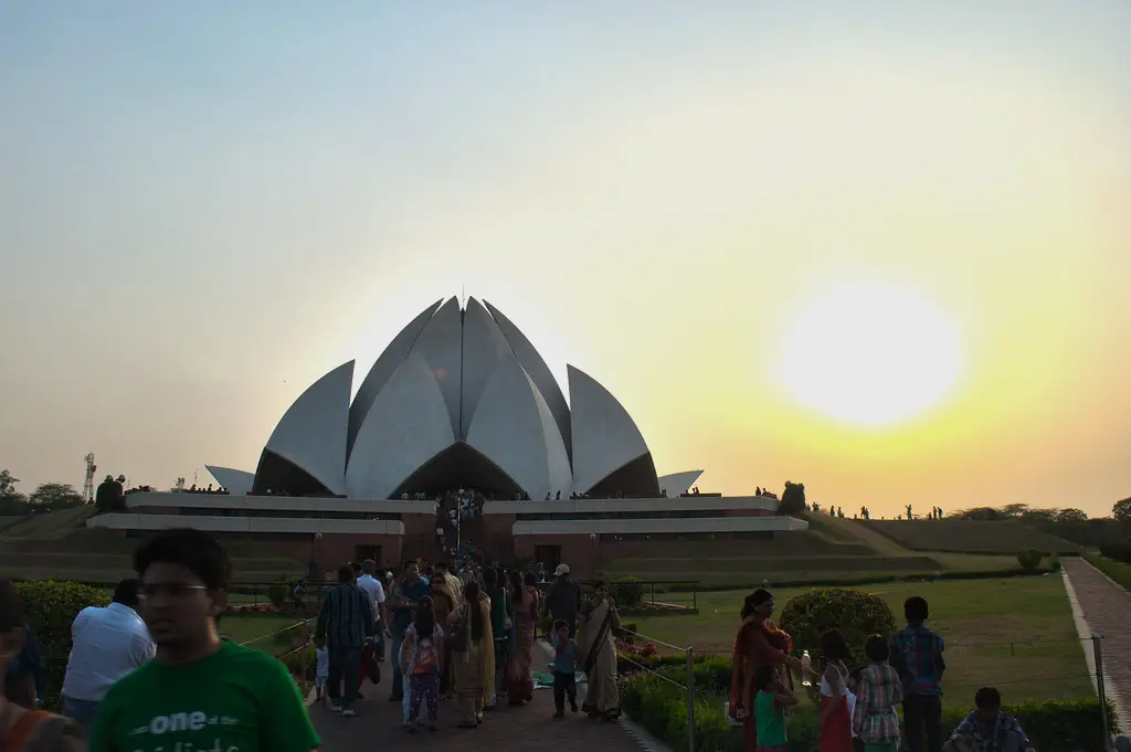 The Lotus Temple, a white, lotus-shaped Baháʼí House of Worship, is silhouetted against a bright, setting sun, with many visitors walking around its grounds in New Delhi, India.