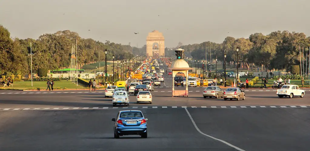 India Gate, the All India War Memorial, stands prominently at the end of the bustling Kartavya Path, which is filled with cars and flanked by green lawns and trees under a hazy sky.