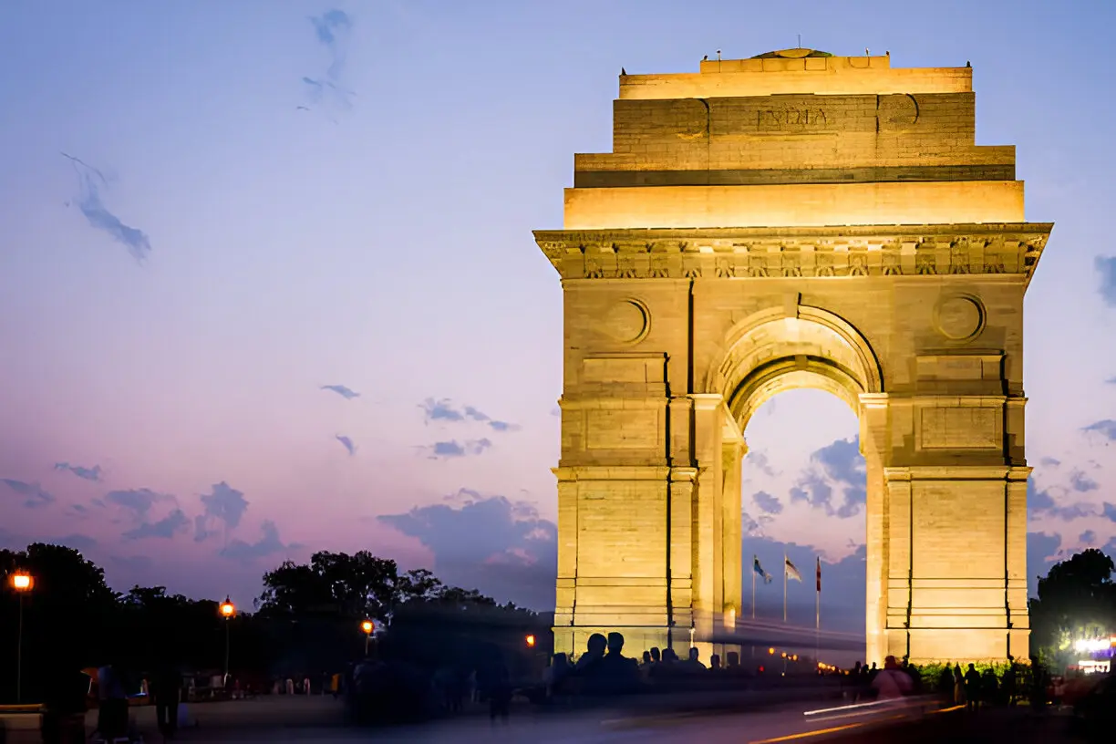 India Gate, a grand archway war memorial, is brightly illuminated in golden light against a twilight sky painted with shades of purple and pink, with light trails from vehicles in the foreground.
