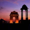 India Gate, an illuminated war memorial arch, stands under a dramatic purple, pink, and orange sunset sky, with a silhouetted canopy in the foreground.