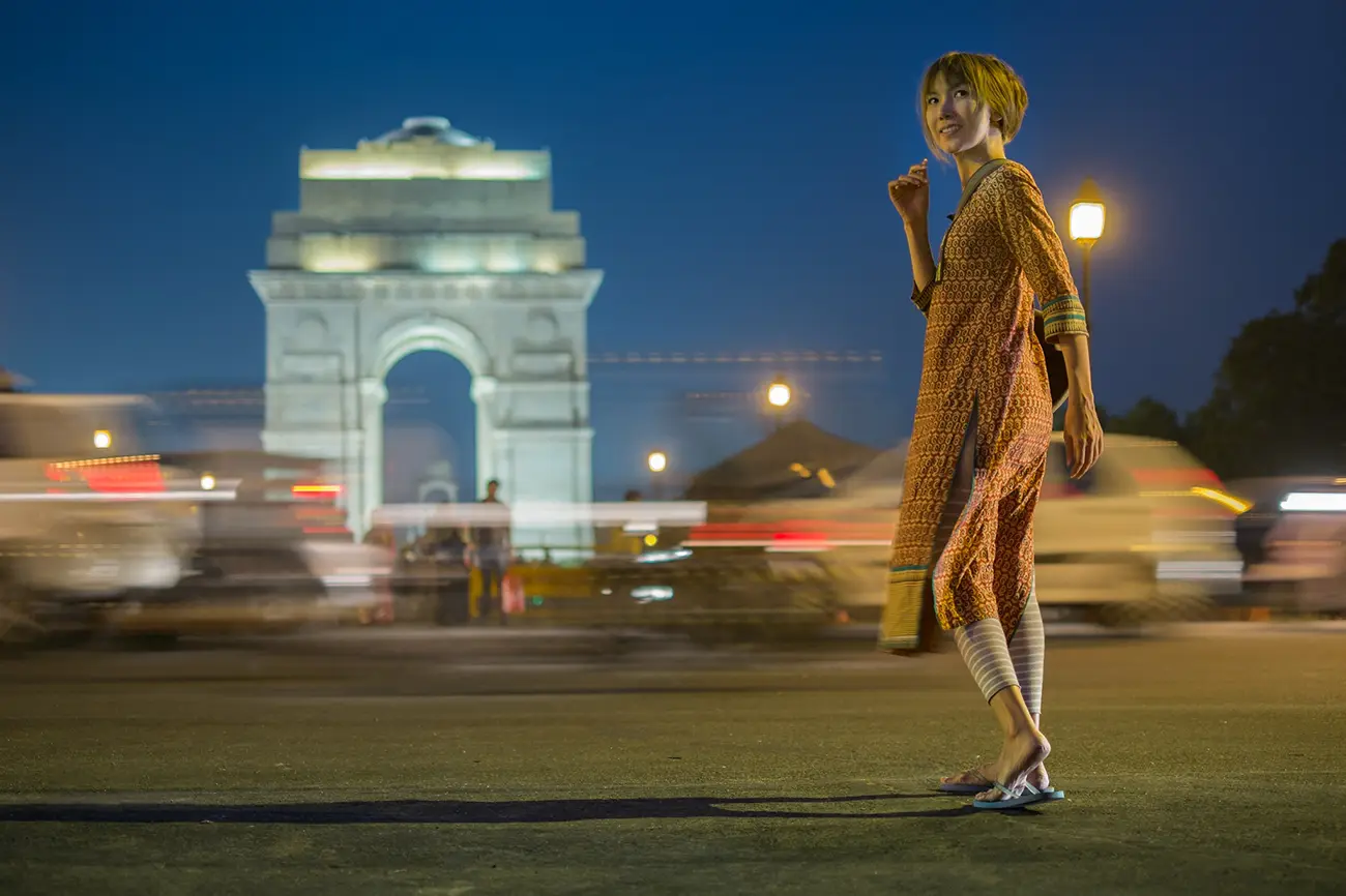 A smiling young woman in a patterned orange tunic and leggings stands in the foreground at night, with the brightly illuminated India Gate and streaks of car lights blurred in the background.