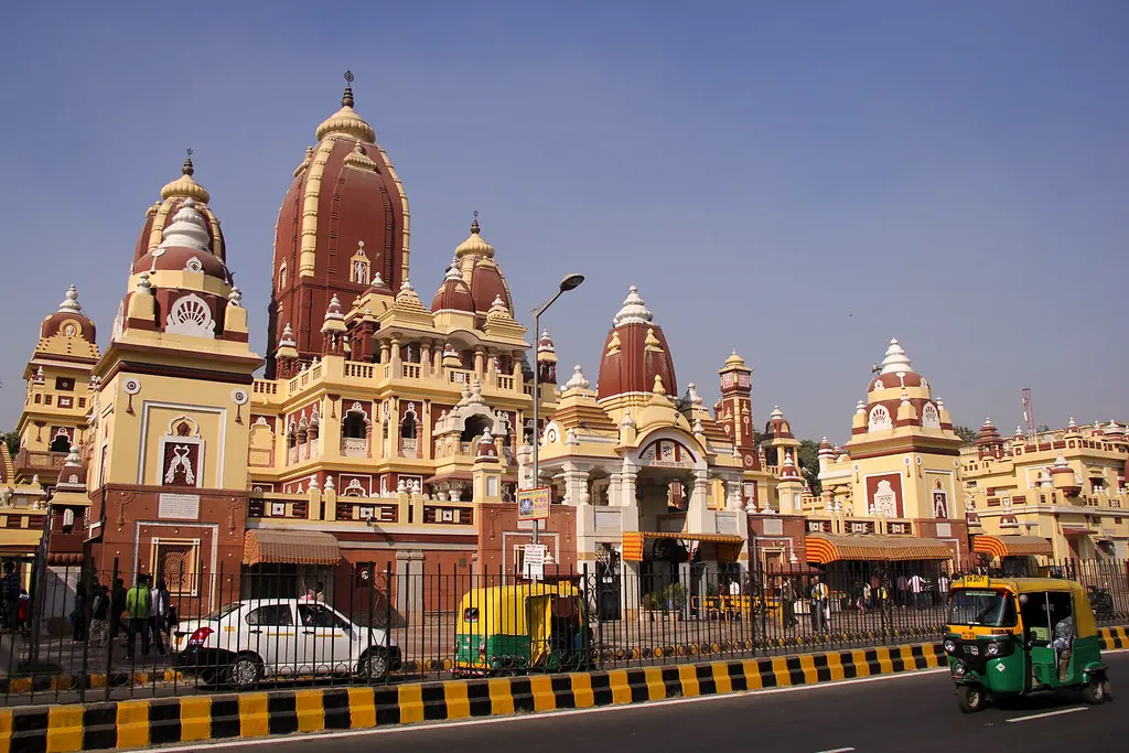 The vibrant Laxmi Narayan Temple (Birla Mandir) in Delhi, with its striking red and cream spires and domes, stands beside a bustling street with traffic and pedestrians under a clear blue sky.