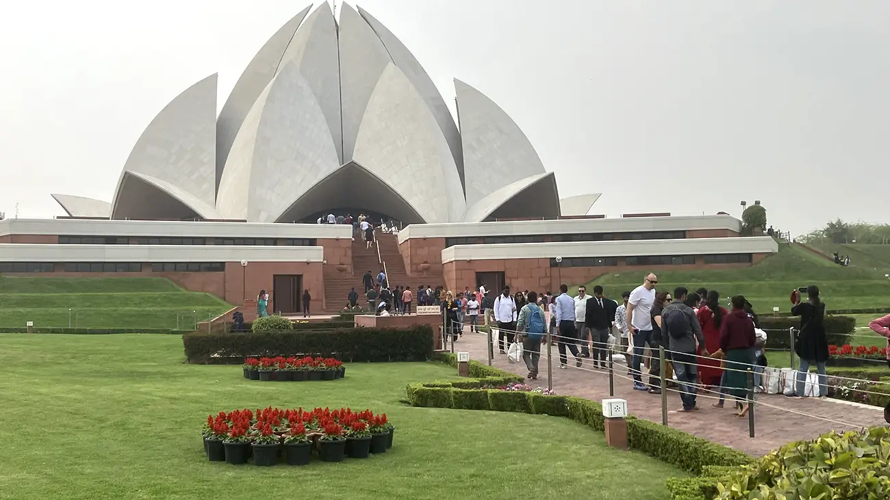 The distinctive white, lotus-shaped Baháʼí House of Worship (Lotus Temple) in Delhi, with many visitors walking on pathways towards its entrance, surrounded by green lawns and flowerbeds.