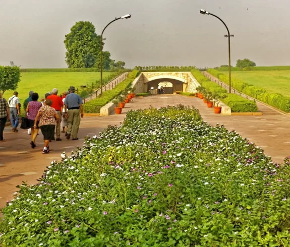 A group of visitors walking along a wide, paved path lined with manicured shrubs and flower beds towards a tunnel-like entrance at Raj Ghat, Delhi, under a clear sky.