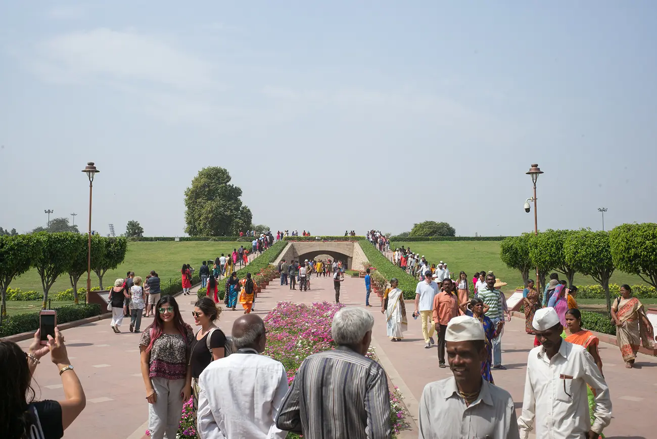 A wide view of Raj Ghat memorial in Delhi, showing a large crowd of diverse visitors walking on the pathways and grassy areas under a clear sky.