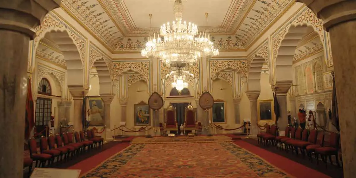 The grand Sabha Niwas, a large hall with ornate white walls and ceiling featuring intricate patterns, multiple chandeliers hanging from above, and rows of red velvet chairs lining a central red carpet leading to two prominent thrones.